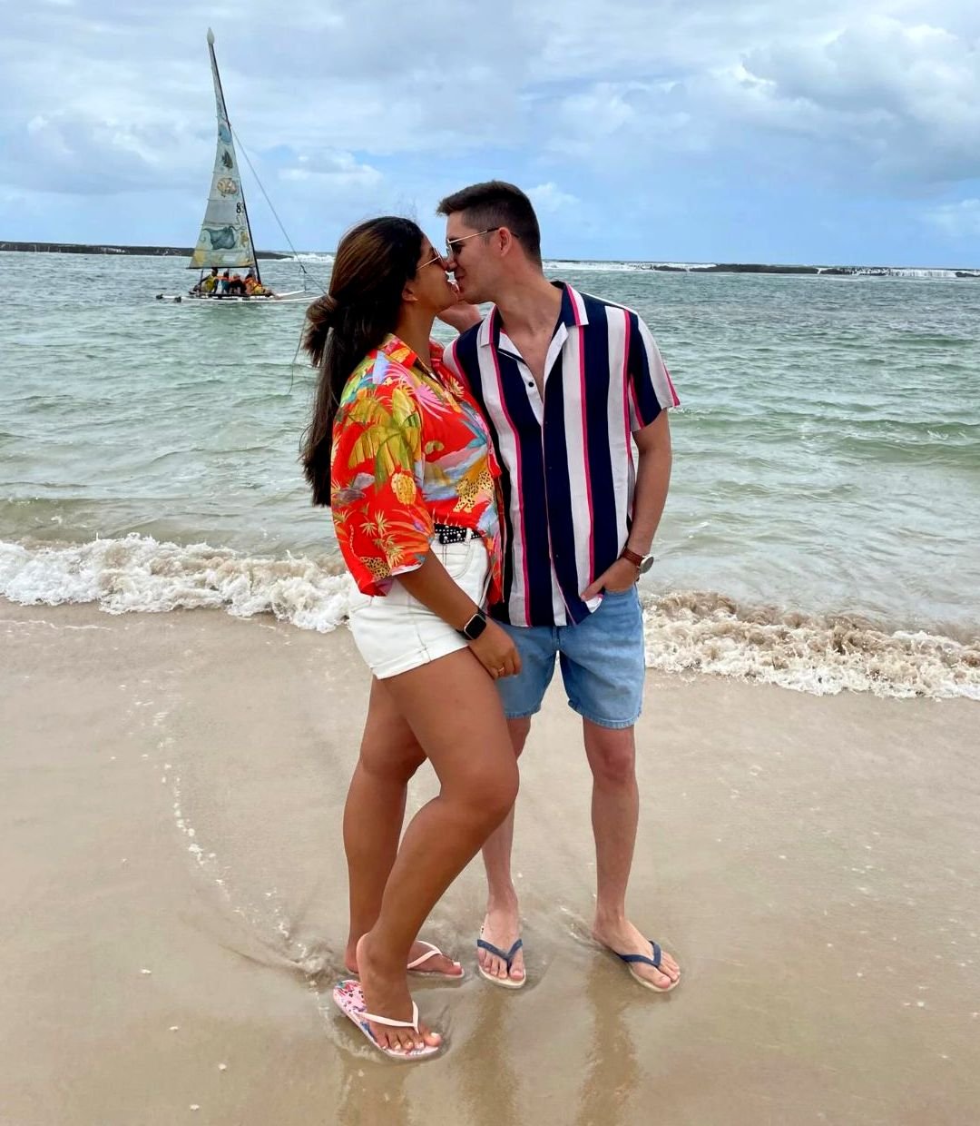 Jessica e Felipe abraçados na praia, com um veleiro ao fundo